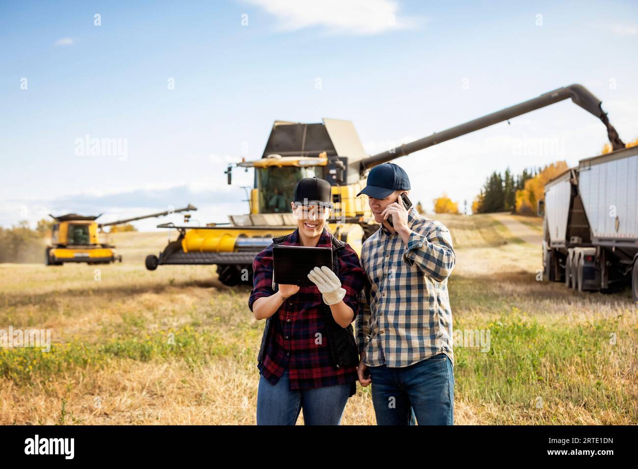 Ehemann und Ehefrau verwenden tragbare drahtlose Geräte zur Verwaltung und Überwachung ihrer Rapsernten und tätigen einen Anruf, während der Mähdrescher Getreide entlädt... Stockfoto