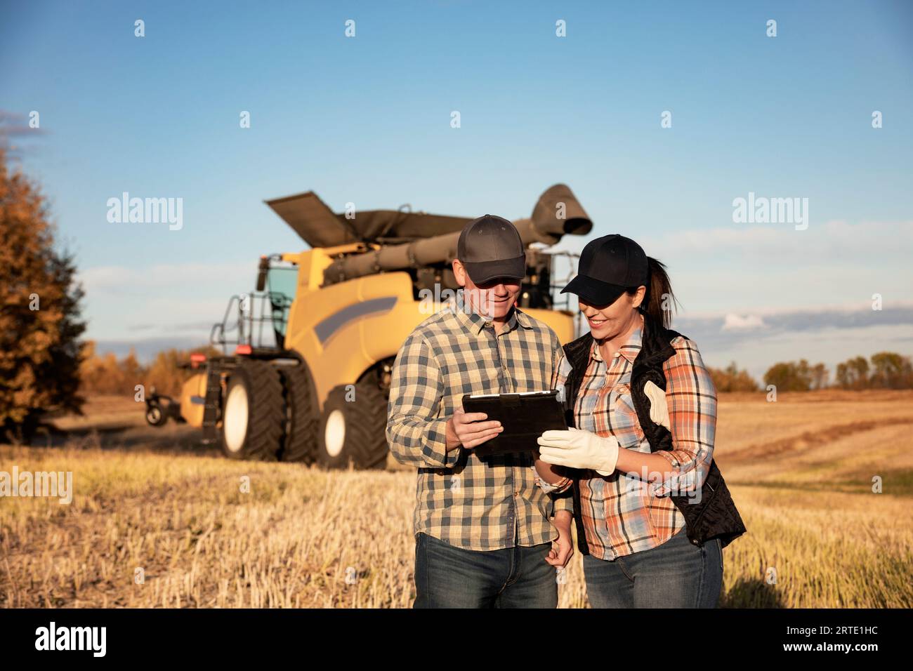 Ein Mann und eine Frau stehen vor einem Mähdrescher und benutzen ein tragbares drahtloses Gerät, um den Ertrag während ihres Sturzes zu verwalten und zu überwachen... Stockfoto