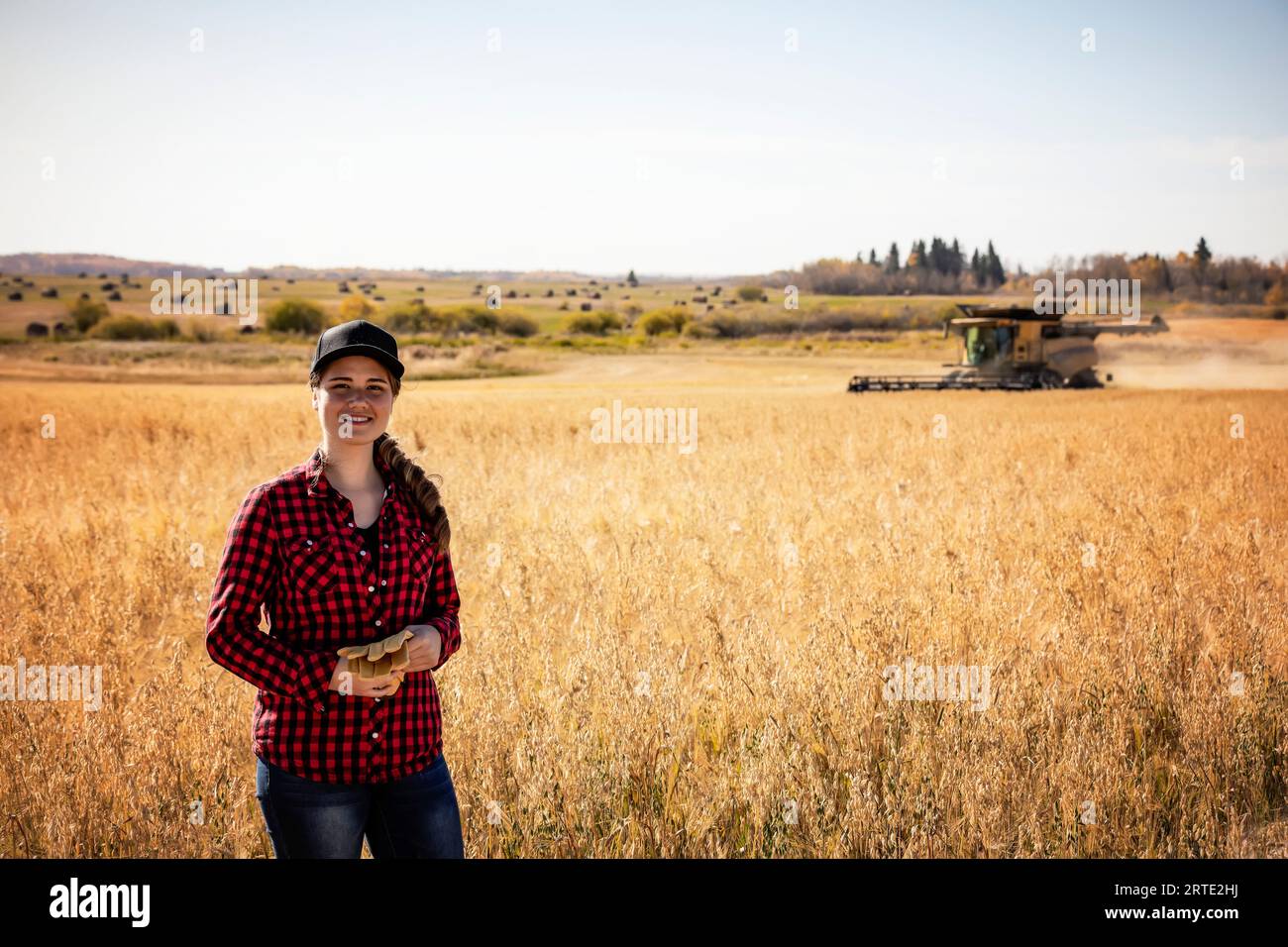 Porträt einer jungen Landwirtschaftsfrau, die zur Erntezeit auf einem Mischkornfeld steht, während ein Mähdrescher im Hintergrund arbeitet Stockfoto