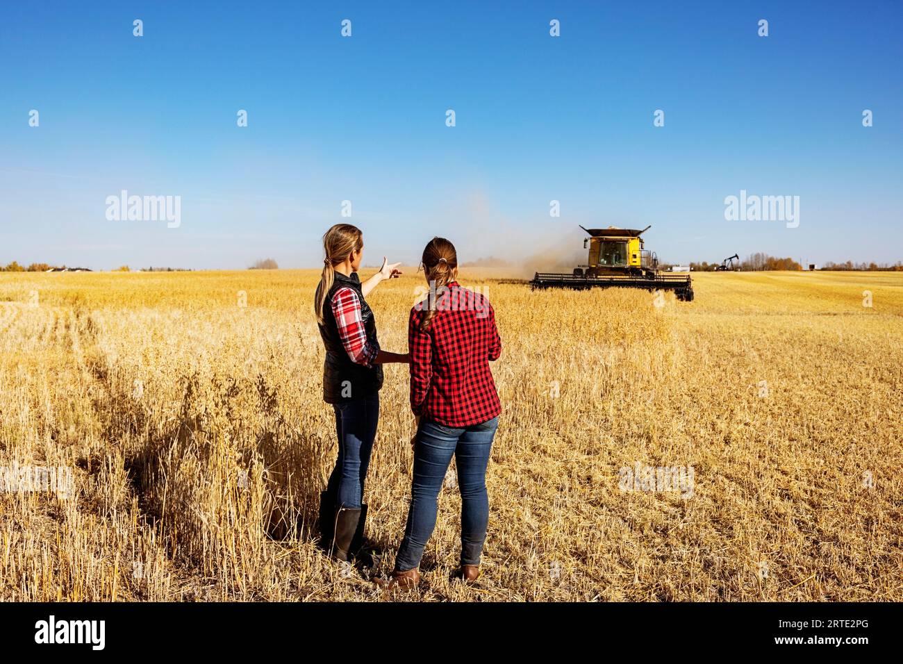 Ein Blick von hinten auf eine reife Landwirtschaftsfrau, die auf einem Feld mit einer jungen Frau zur Erntezeit arbeitet, während ein Mähdrescher in der... Stockfoto