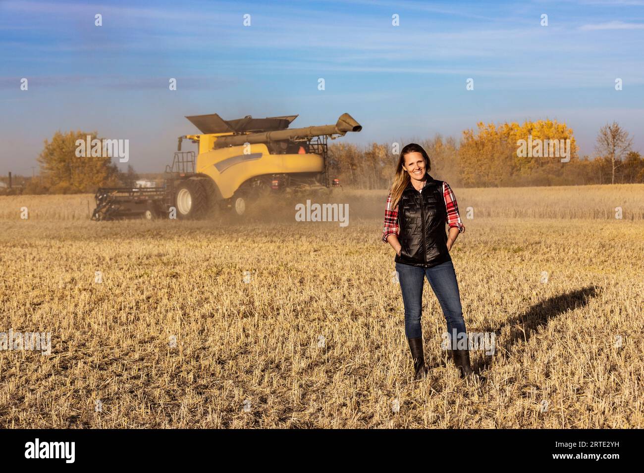 Porträt einer Reifen Landwirtschaftsfrau, die auf einem Getreidefeld steht und während der Ernte für die Kamera posiert, während ein Mähdrescher bei Sonnenuntergang im Hintergrund arbeitet Stockfoto