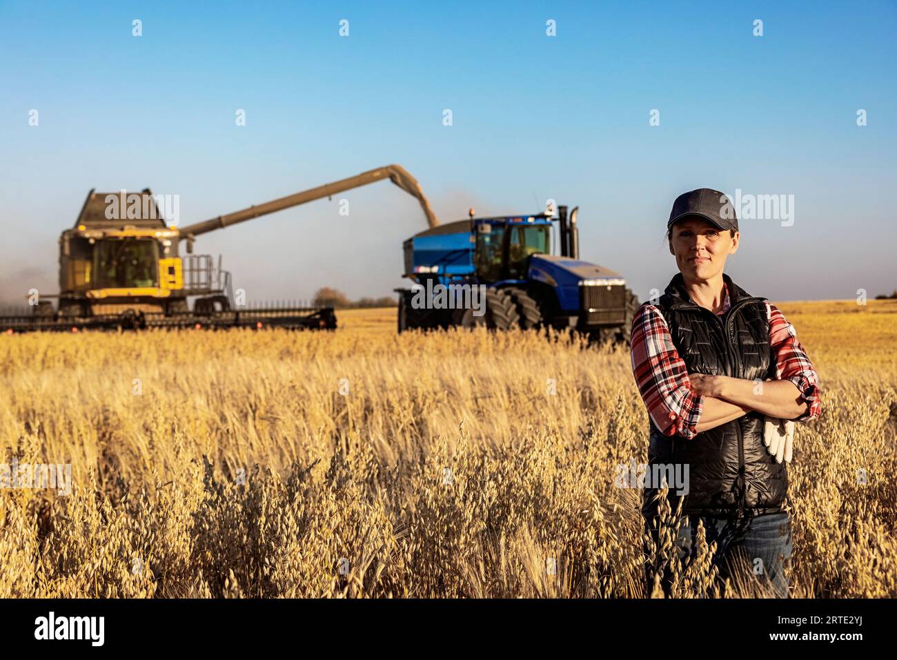 Porträt einer Reifen Landwirtschaftsfrau, die auf einem Getreidefeld steht und während der Ernte für die Kamera posiert, während ein Mähdrescher ein Mischgut auf ein Korn ablädt. Stockfoto