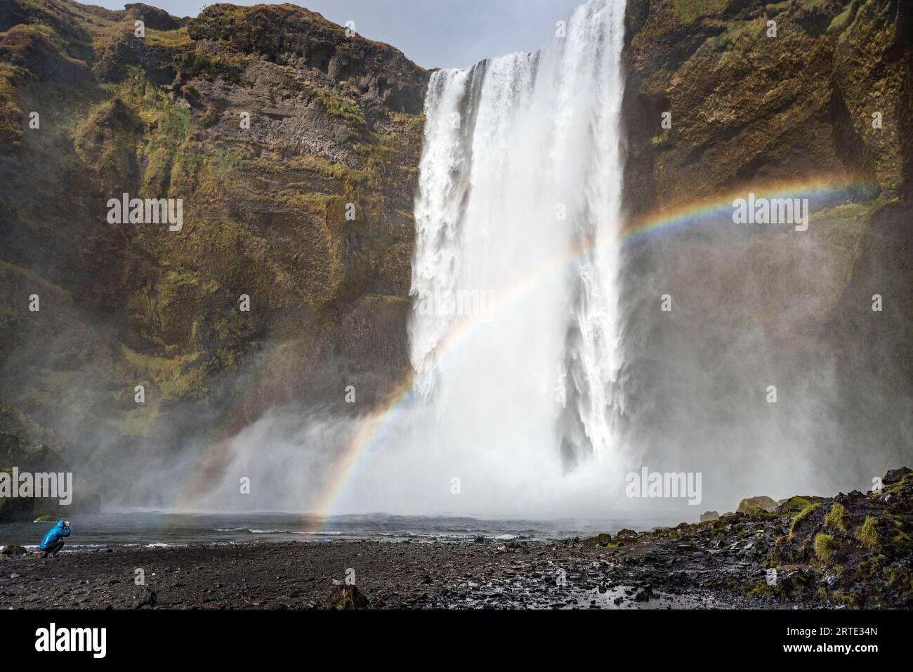 Fotograf, der ein Foto von einem stürzenden Wasserfall und Regenbogen macht; Island Stockfoto