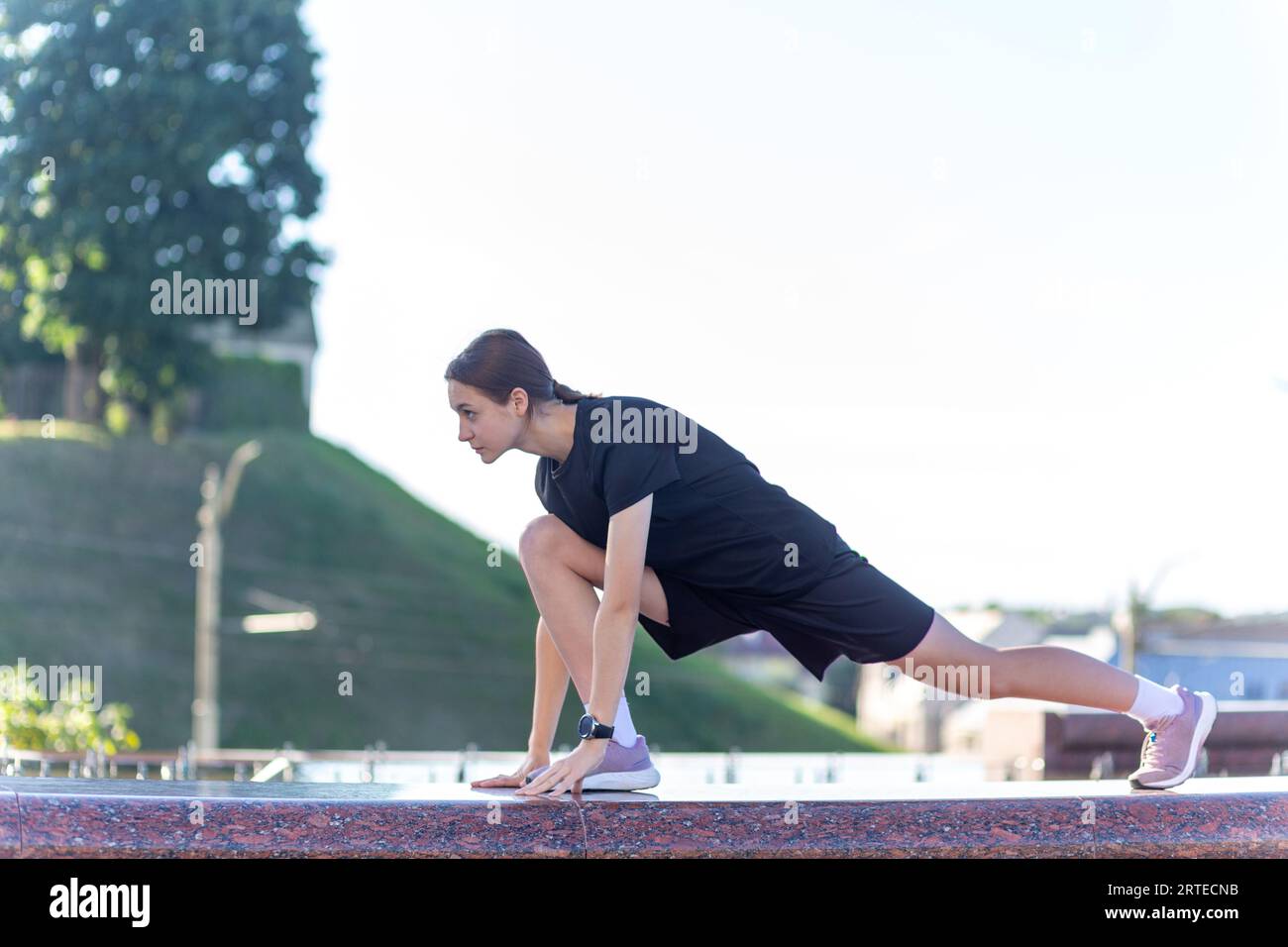Ein junges, sportliches Mädchen in schwarzer Kleidung, das sich nach dem Workout in der Nähe des öffentlichen Brunnens im Stadtpark ausdehnt. Fitness, Sport, Urban Joggin Stockfoto