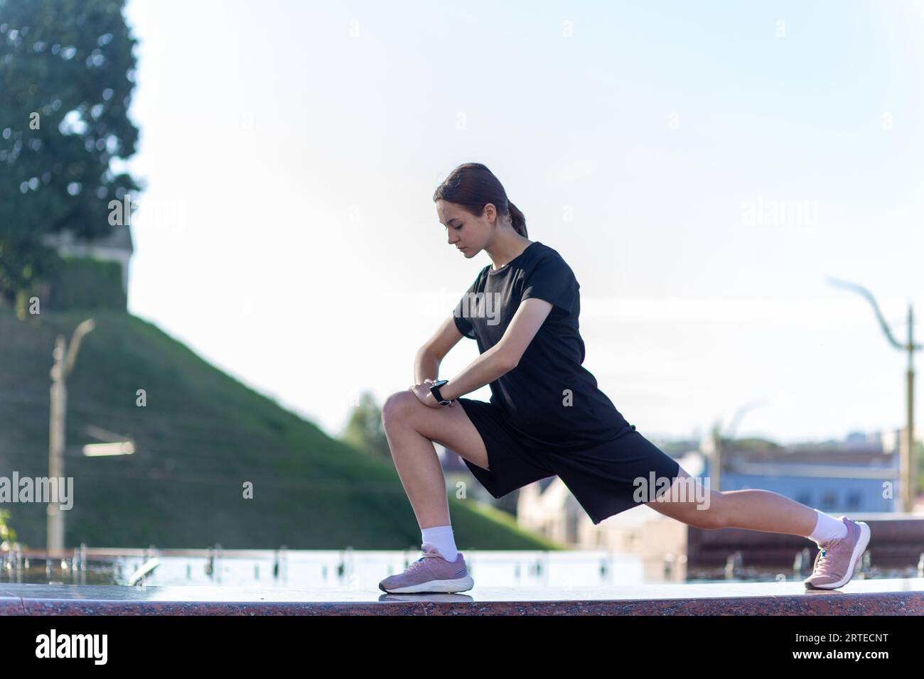 Ein junges, sportliches Mädchen in schwarzer Kleidung, das sich nach dem Workout in der Nähe des öffentlichen Brunnens im Stadtpark ausdehnt. Fitness, Sport, Urban Joggin Stockfoto