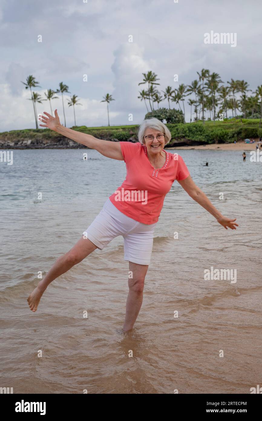 Porträt einer Reifen Frau, die in verspielter Position im Wasser steht und am Sandstrand des Kapalua Resort in West Maui in die Kamera lächelt Stockfoto