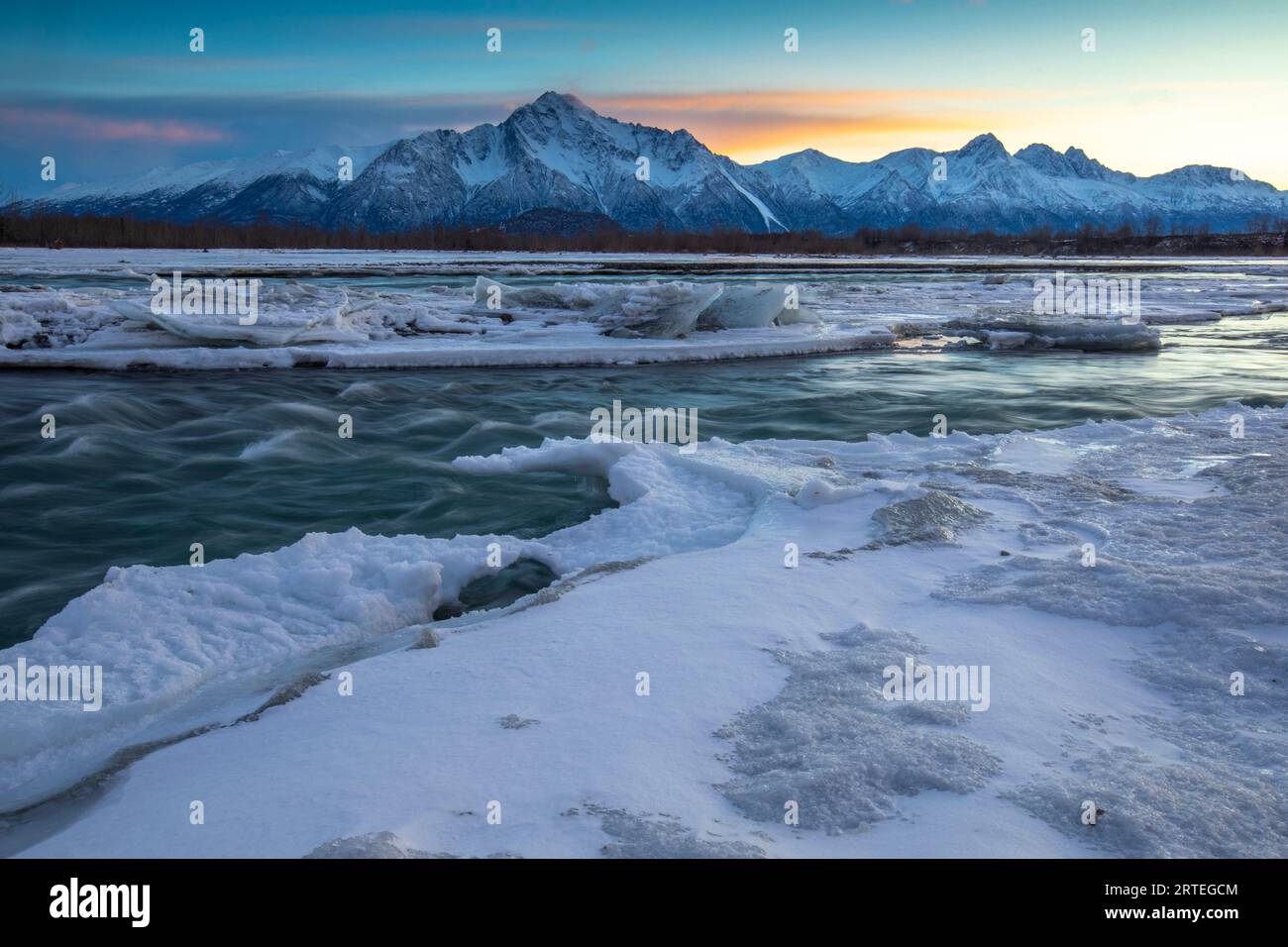 Die Sonne geht hinter Pioneer und Twin Peaks und der Chugach Mountain Range in einer Winternacht unter, mit einem seidenglatten Matanuska River, gesäumt von Eis... Stockfoto