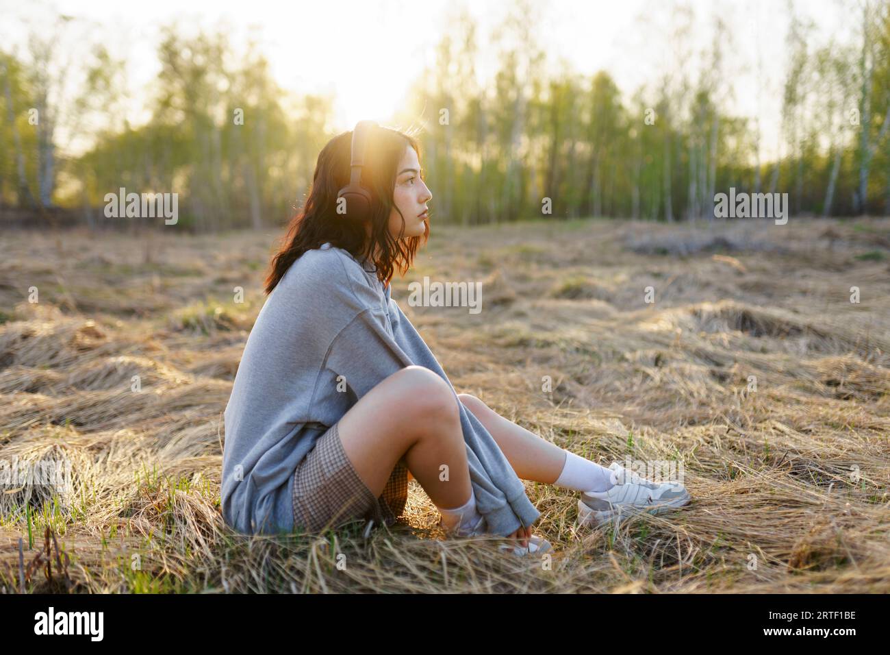 Seitenansicht einer Frau, die Musik hört, während sie bei Sonnenuntergang auf Gras auf der Wiese sitzt Stockfoto