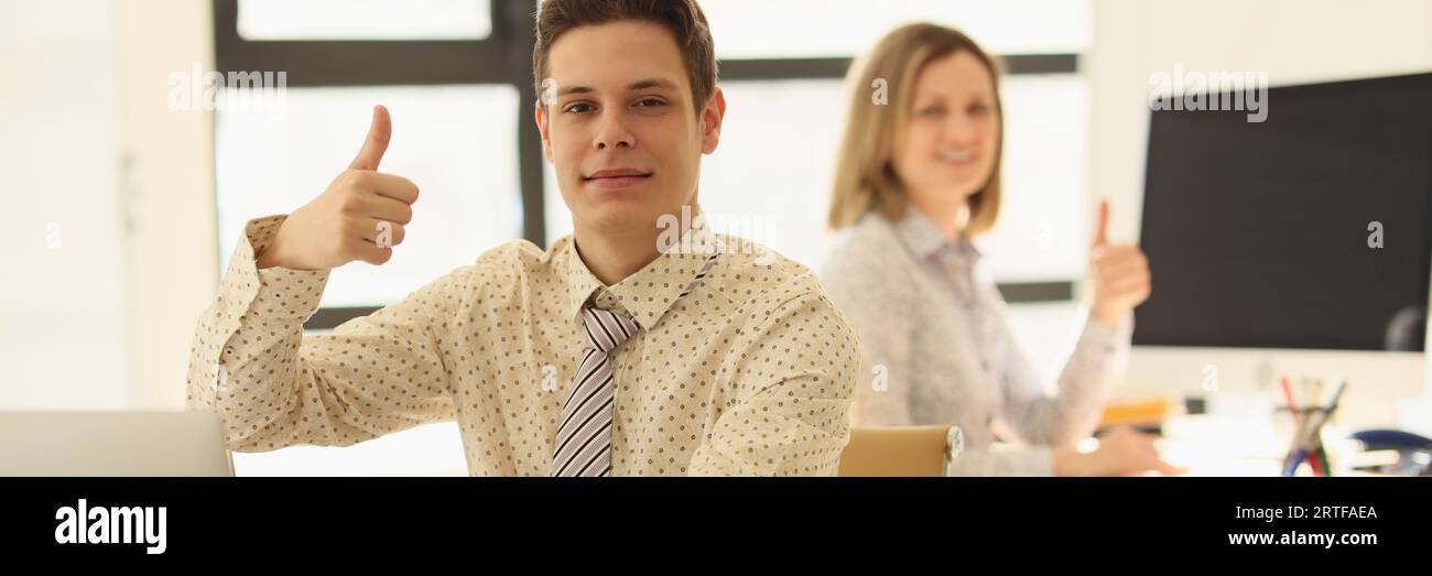 Zwei Manager sitzen im Büro an ihrem Schreibtisch und zeigen Daumen-hoch-Geste. Stockfoto