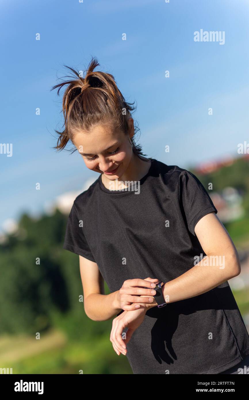 Teenager-Mädchen in schwarzer Kleidung, das nach einem Workout ihre Fitnessuhr überprüft. Müde Läuferin, die die Pulsationen kontrolliert und die Smartwatch überprüft. Stockfoto