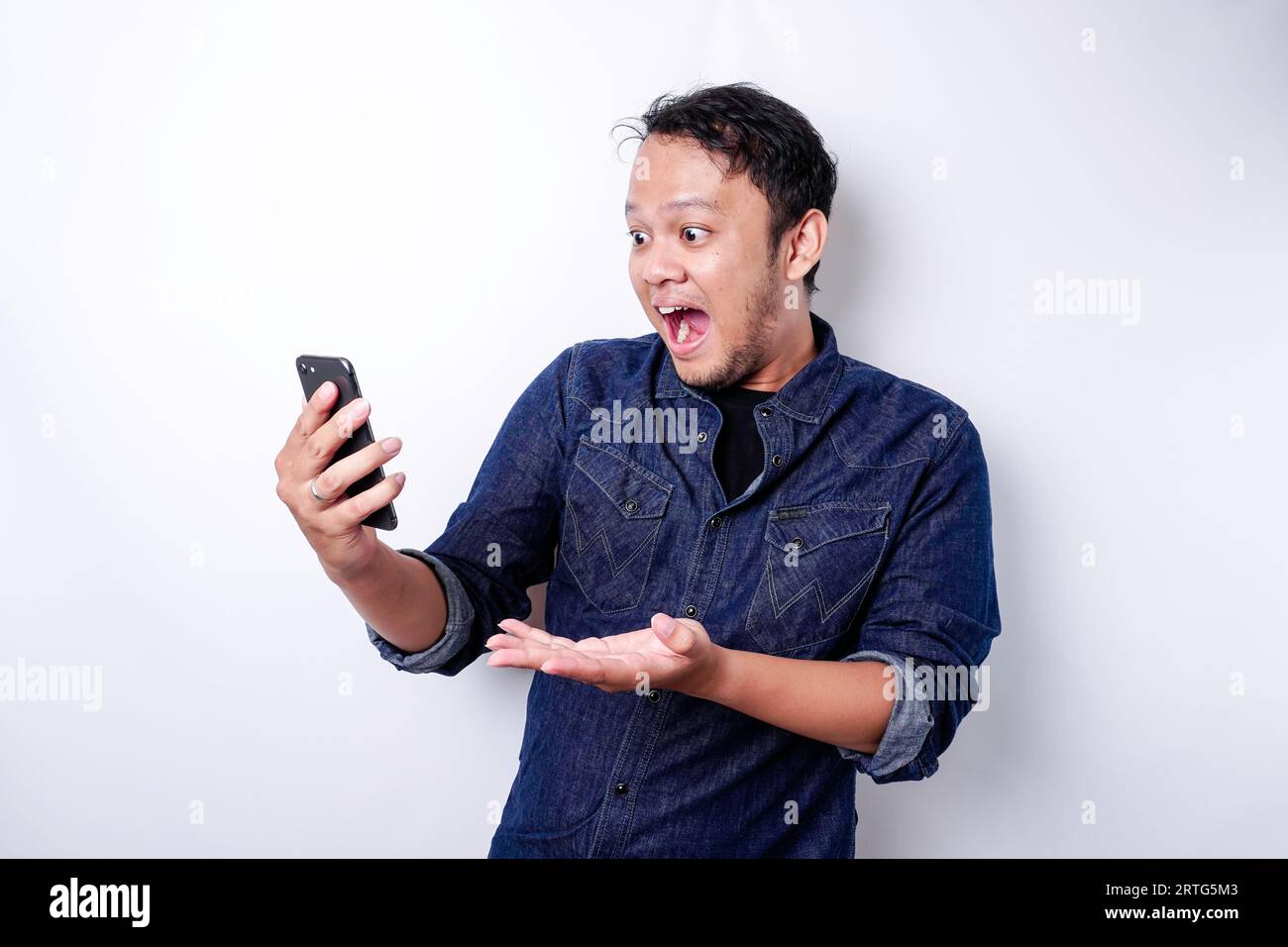 Schockierter asiatischer Mann, der blaues Hemd trägt und sein Telefon hält, isoliert durch weißen Hintergrund Stockfoto