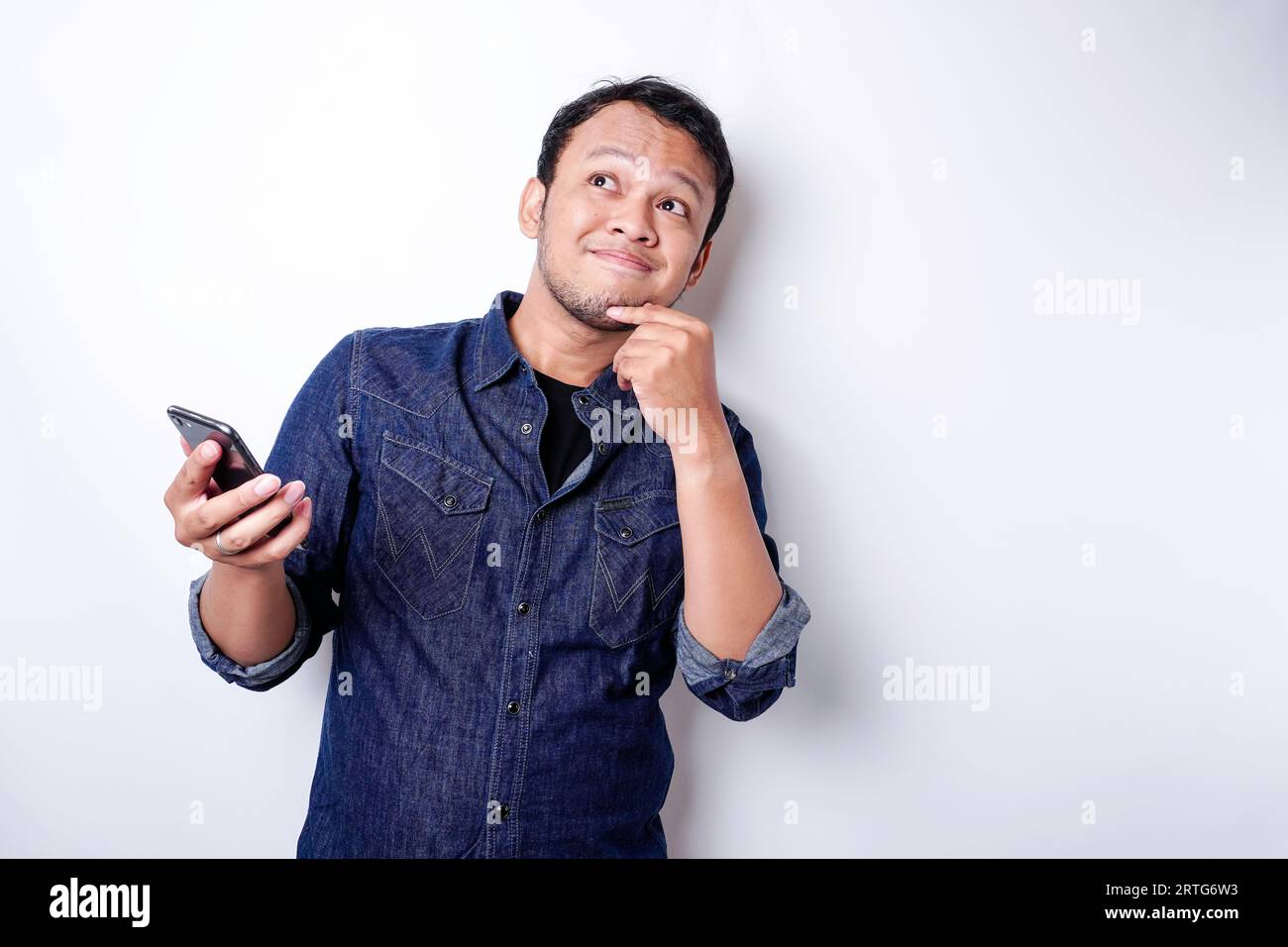 Ein nachdenklicher junger Mann, der in einem blauen Hemd gekleidet ist, während er sein Telefon mit dem weißen Hintergrund abhält Stockfoto