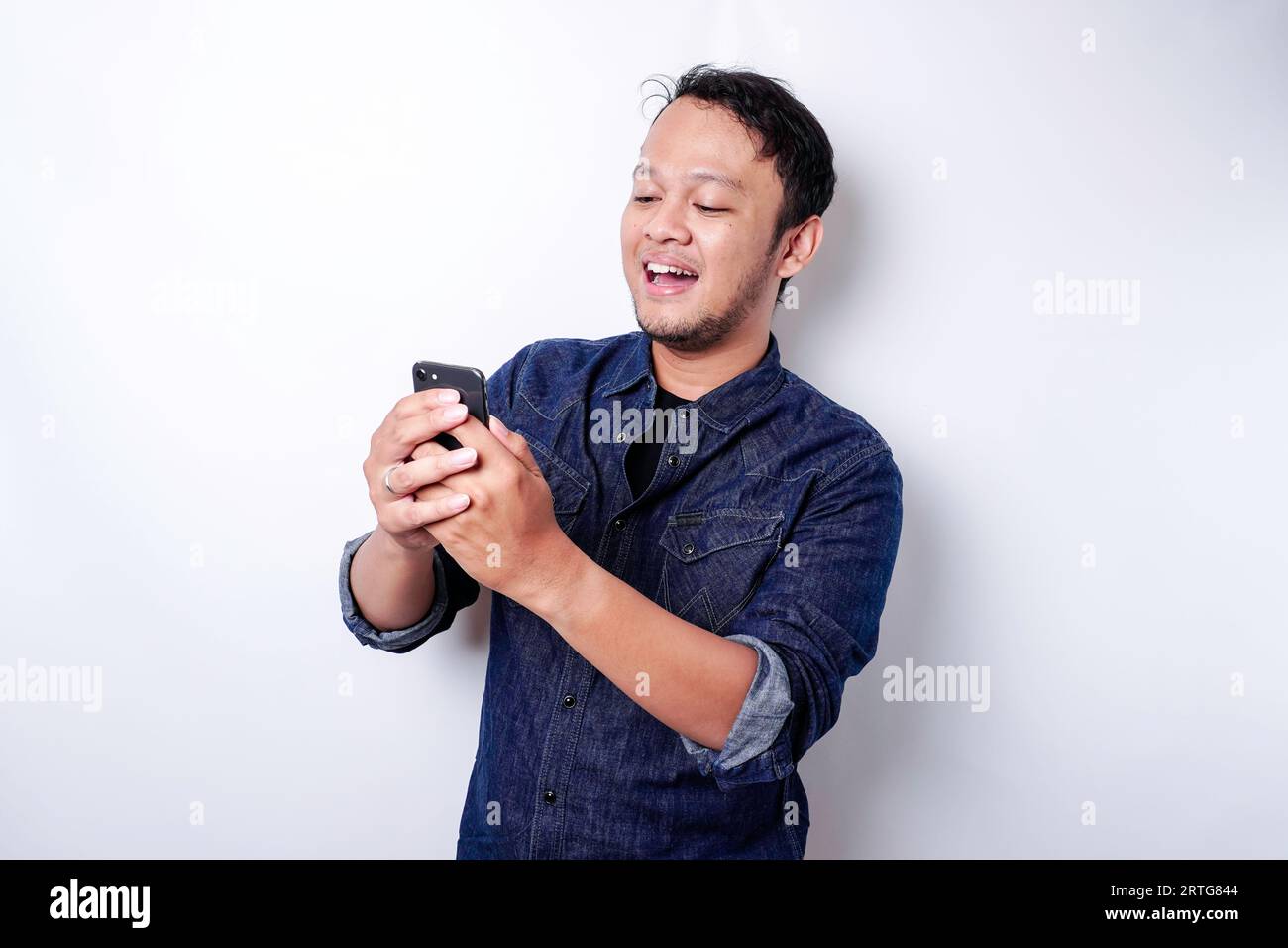 Aufgeregter asiatischer Mann, der blaues Hemd trägt und lächelt, während er sein Telefon hält, isoliert durch weißen Hintergrund Stockfoto