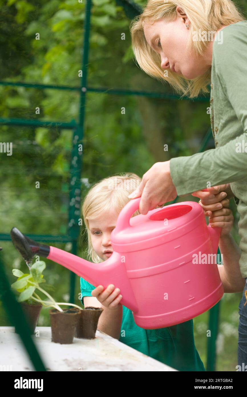 Junge blonde Mädchen hilft Frau Sämling Töpfe im Gewächshaus mit rosa Gießkanne gießen Stockfoto