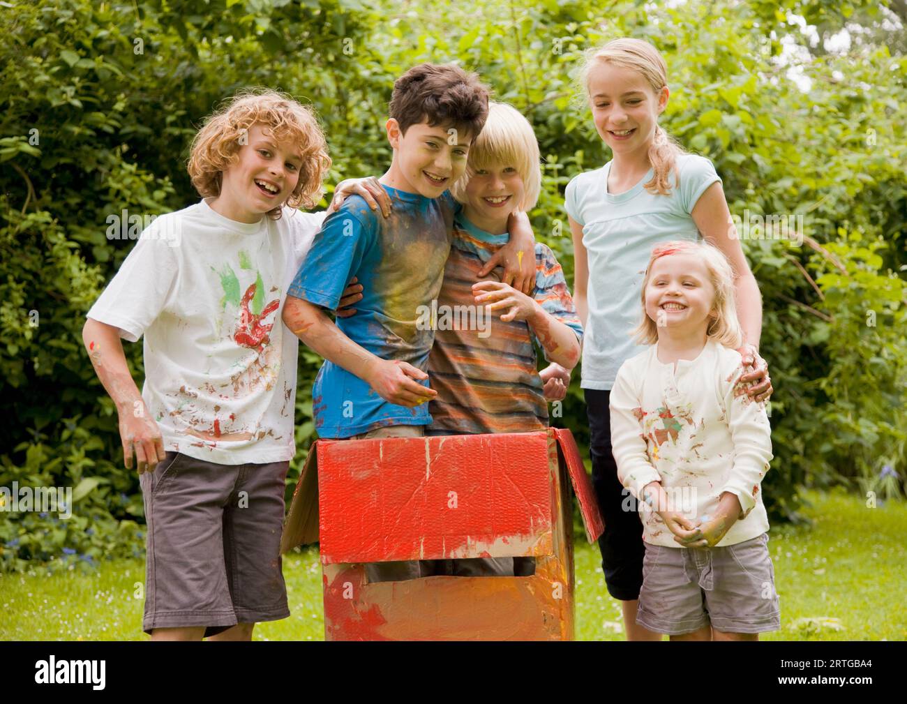 Kinder in einem Garten mit Aquarellfarbe bedeckt, zwei von ihnen stehen in einer Pappschachtel Stockfoto