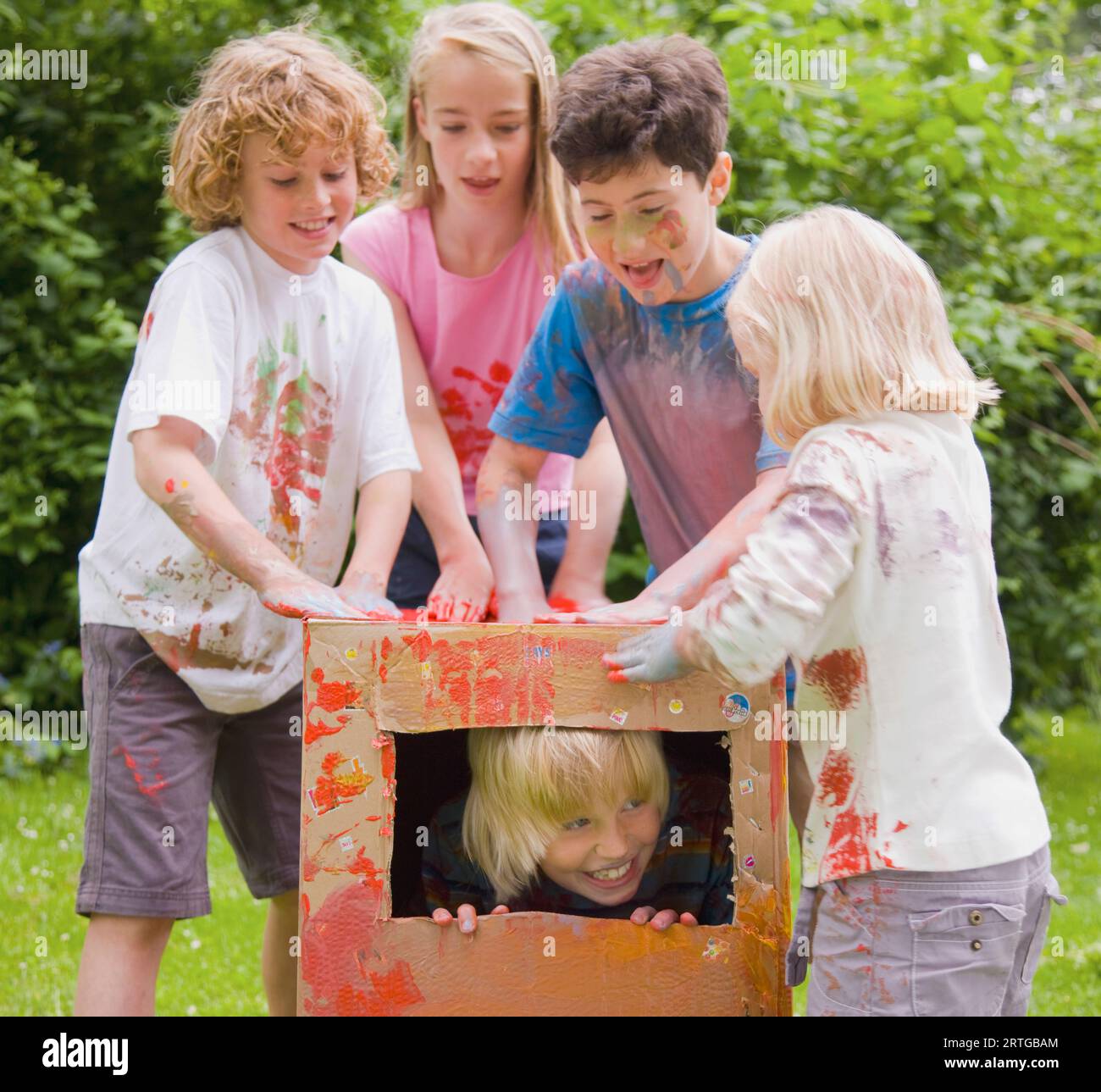 Kinder, die mit Aquarellfarbe bedeckt sind und im Garten spielen, eines davon in einer Pappschachtel Stockfoto