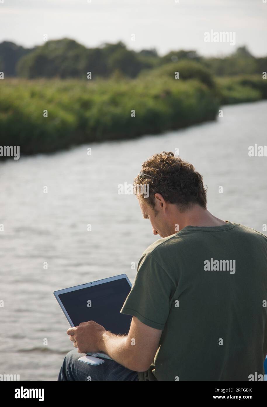 Rückansicht des ein Mann sitzt an einem Fluss mit einem Laptopcomputer Stockfoto