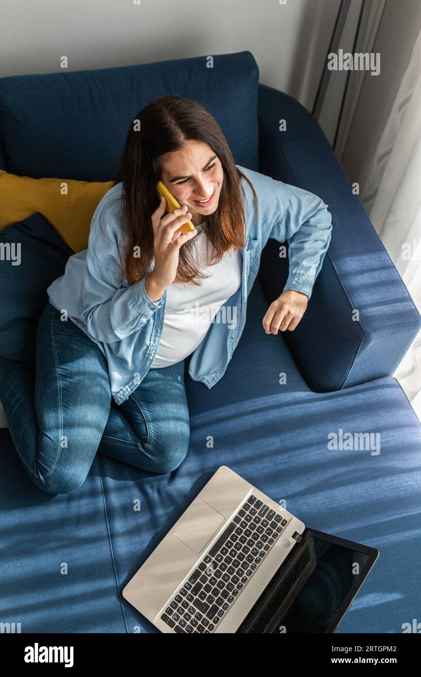 Schwangerin sitzt auf dem Sofa mit Laptop im Wohnzimmer Zimmer und sprechen auf dem Handy, während Sie von zu Hause aus arbeiten Stockfoto