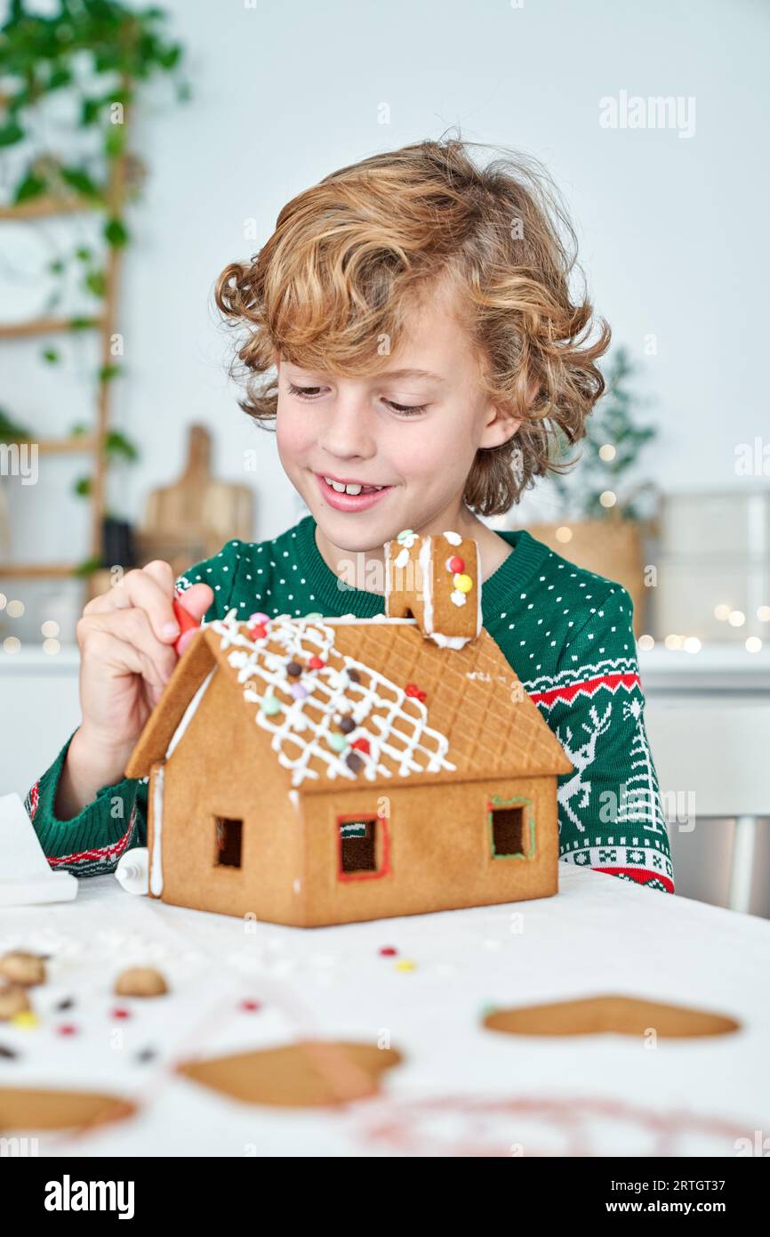 Positive Boy-Klebe-Details von Lebkuchenhaus mit süßer Zuckerglasur beim Sitzen am Tisch in der hellen Küche während der Weihnachtsvorbereitung Stockfoto