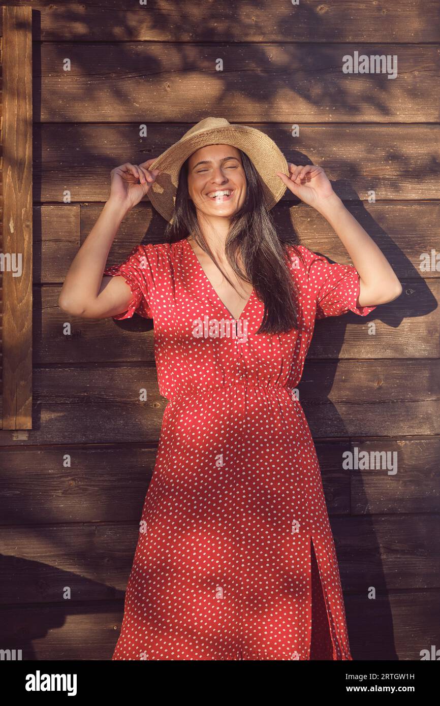 Positives junges Weibchen in rotem Polka-Dot-Kleid und Hut lächelnd mit geschlossenen Augen, während es an sonnigen Tagen auf hölzernem Hintergrund steht Stockfoto