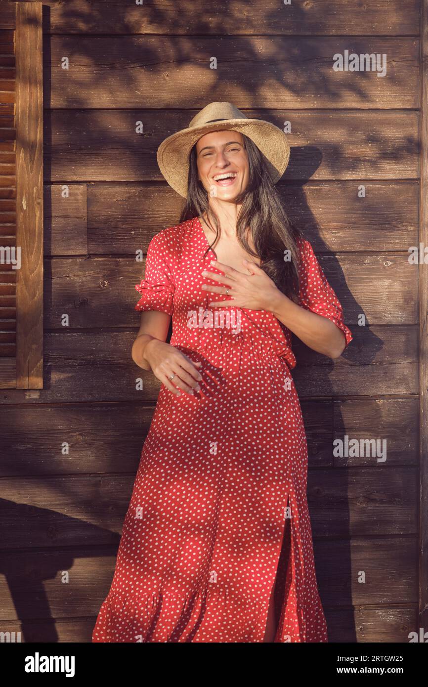 Lachende Frau mit rotem Polka-Dot-Kleid und Strohhut, die Kamera anschaut, während sie an sonnigen Tagen gegen ein Holzhaus steht Stockfoto