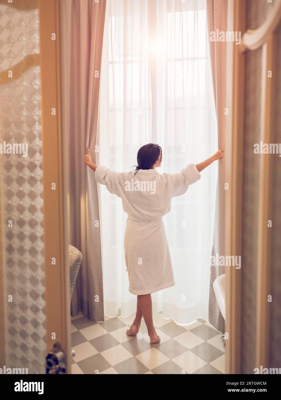 Rückansicht einer anonymen Barfußfrau im Bademantel, die in der Nähe des Fensters steht und die Vorhänge im Hotelbad öffnet, nachdem sie morgens aufwacht Stockfoto