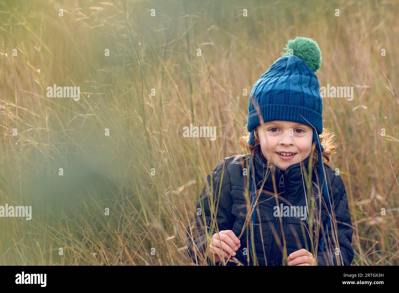 Positiver Junge in warmer Mütze und Oberbekleidung, der mit einem Lächeln auf die Kamera schaut, während er inmitten von hohem, trockenem Gras in der Natur steht Stockfoto