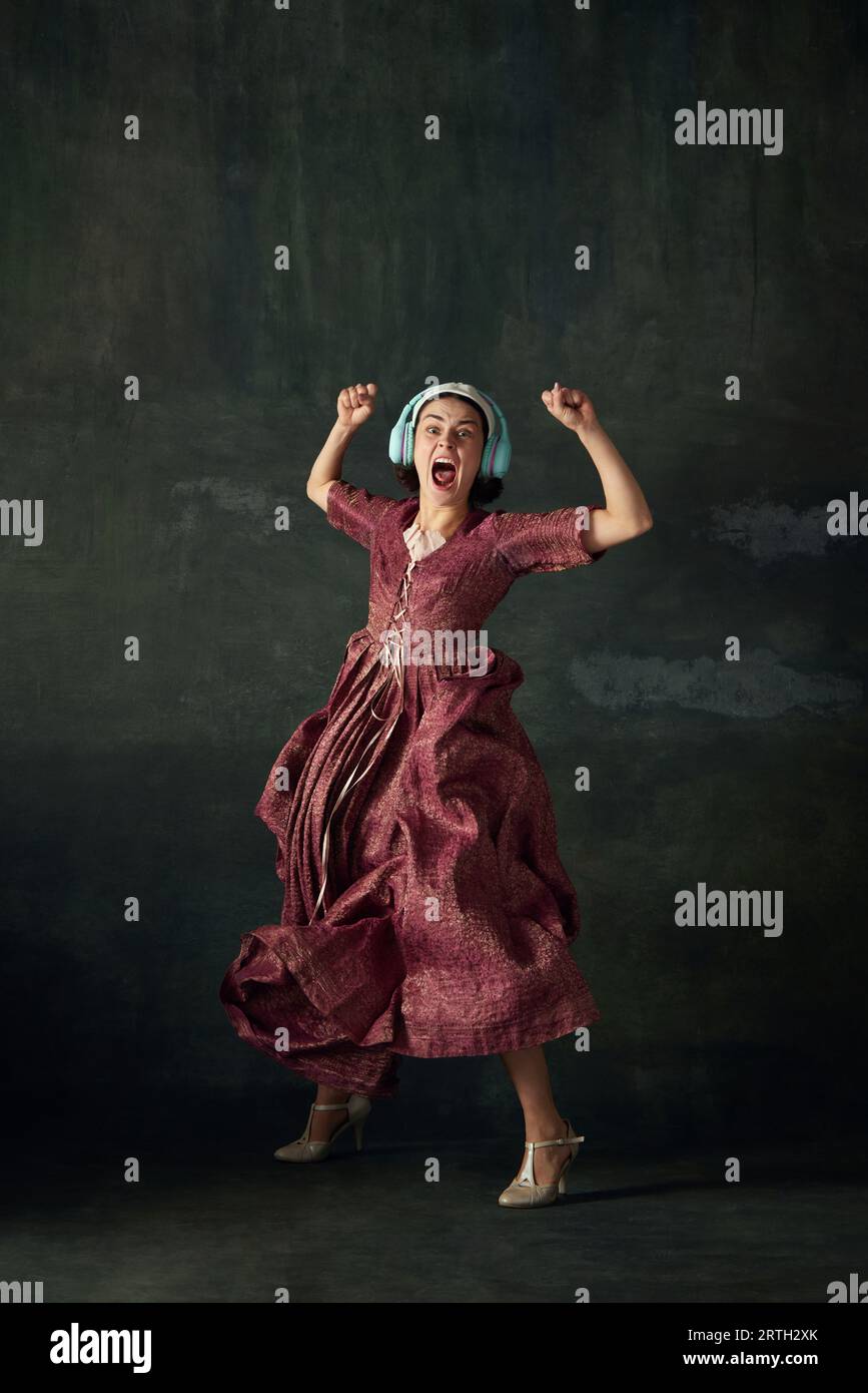 Emotionale Frau in elegantem Renaissance-Kleid, mittelalterliche Dienstmädchen, die Musik im Kopfhörer hört und vor altem grünen Hintergrund tanzt Stockfoto