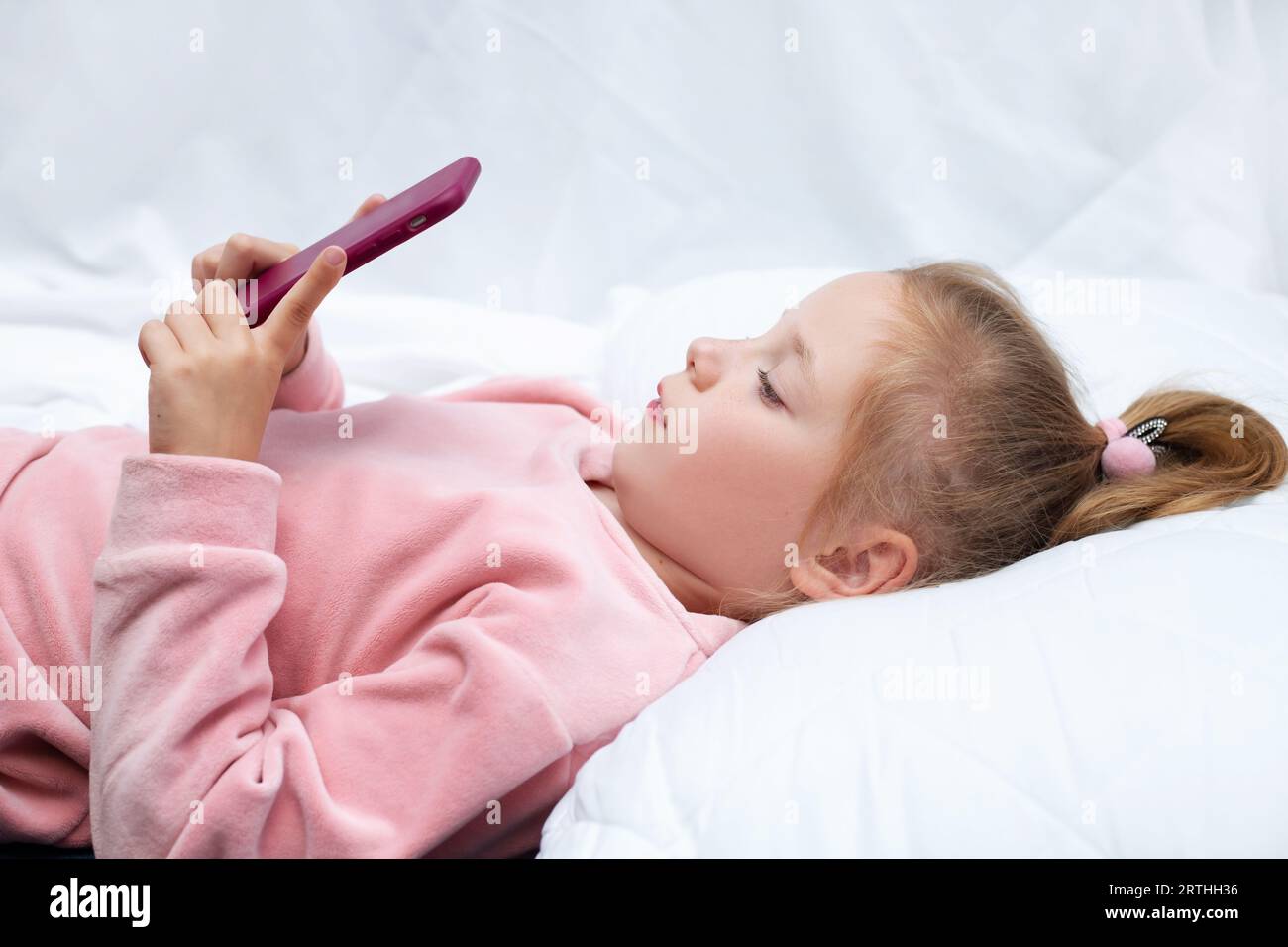 Kleines Mädchen (3-4 Jahre) liegt im Bett und schaut auf den Smartphone-Bildschirm. Das Kind schaut sich Karikaturen an, spielt Spiele vor einem Traum. Stockfoto