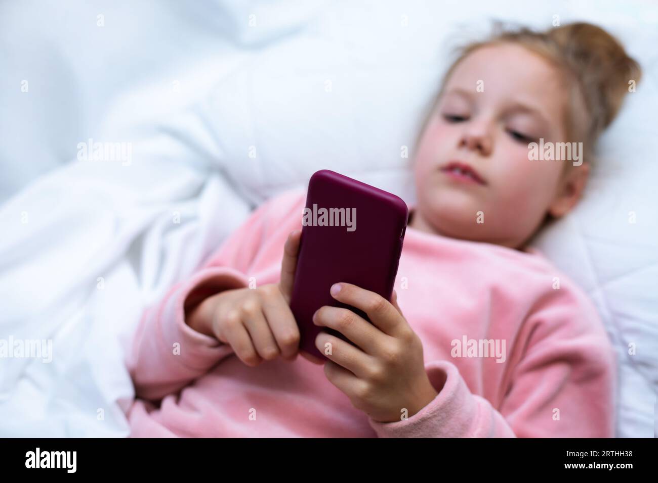 Kleines Mädchen (3-4 Jahre) liegt im Bett und schaut auf den Smartphone-Bildschirm. Das Kind schaut sich Karikaturen an, spielt Spiele vor einem Traum. Stockfoto