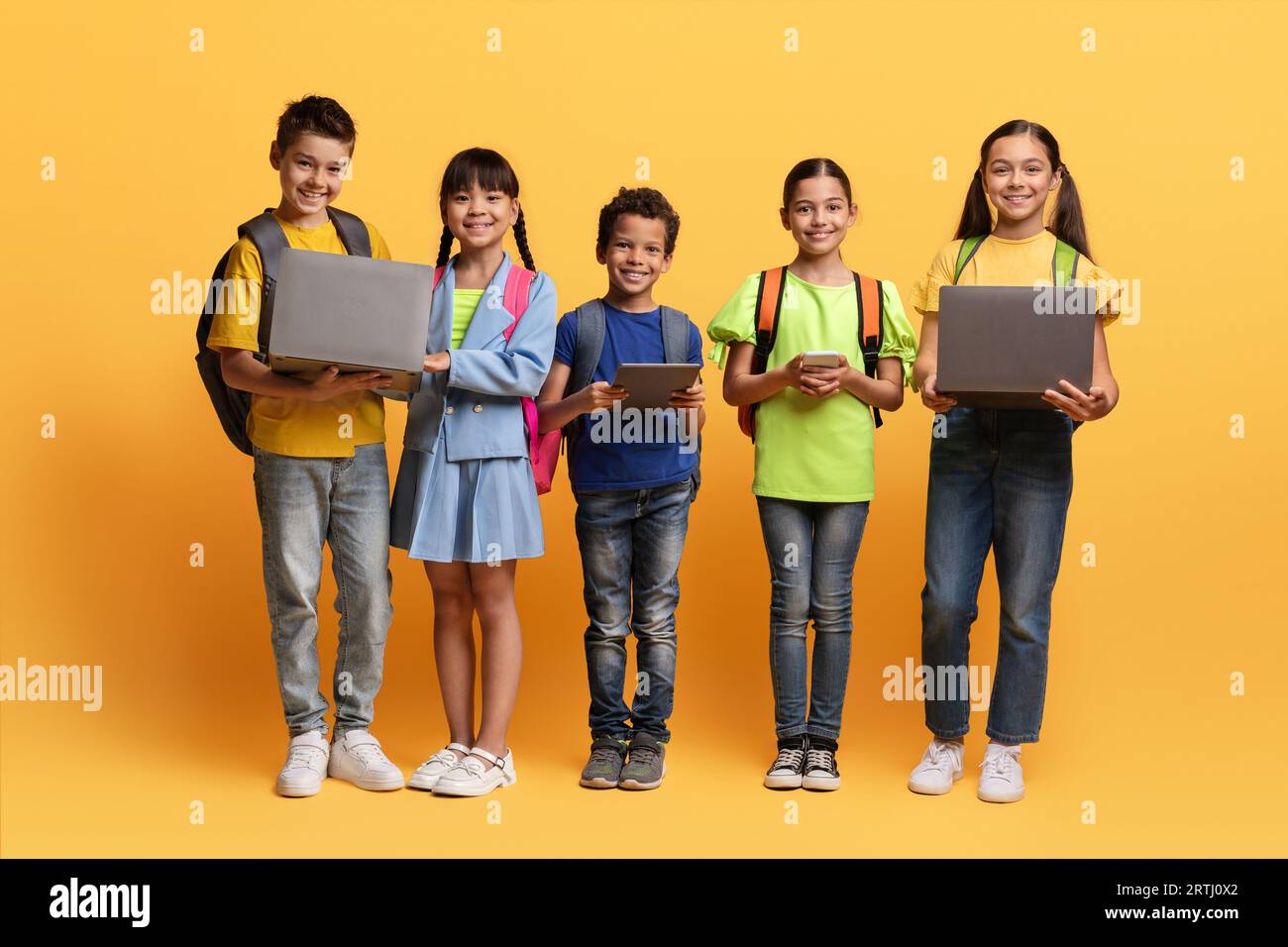 Niedliche multiethnische Schulkinder mit verschiedenen Gadgets auf gelbem Hintergrund Stockfoto