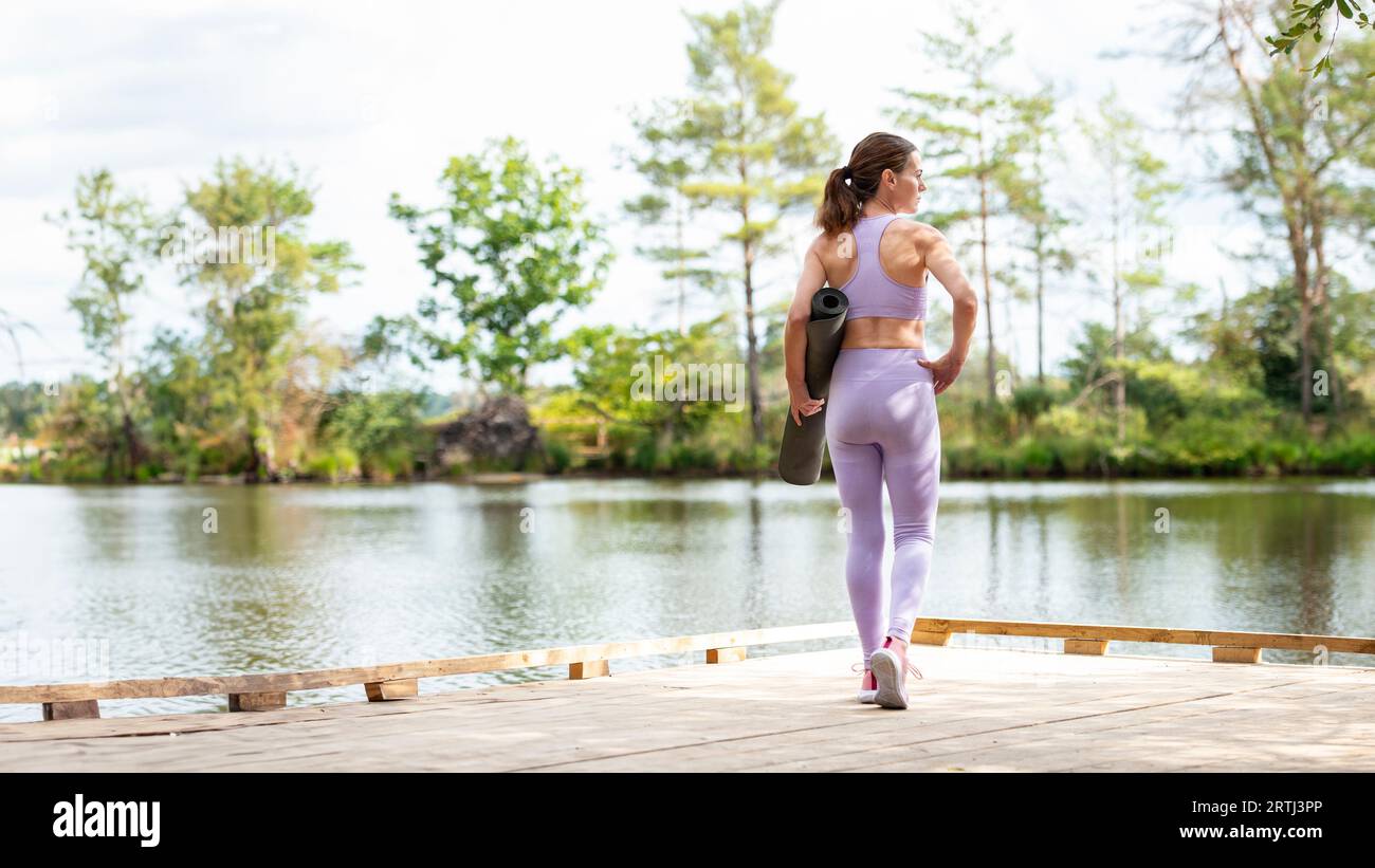 Sportliche Frau, die auf einem Steg am See steht und eine Trainingsmatte hält, Teil der Serie. Stockfoto