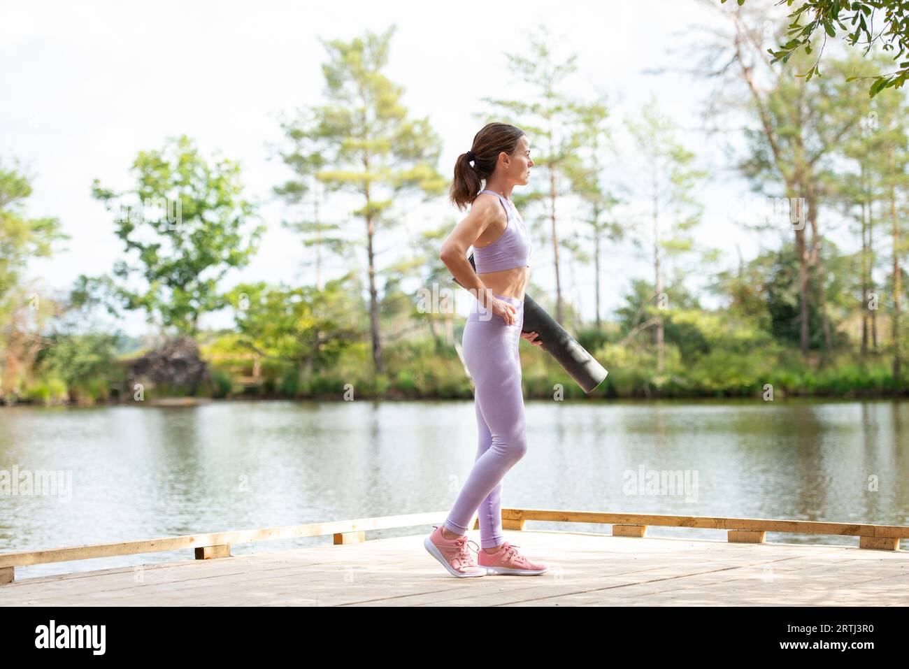 Sportliche Frau, die auf einem Steg am See steht und eine Trainingsmatte hält, Teil der Serie. Stockfoto