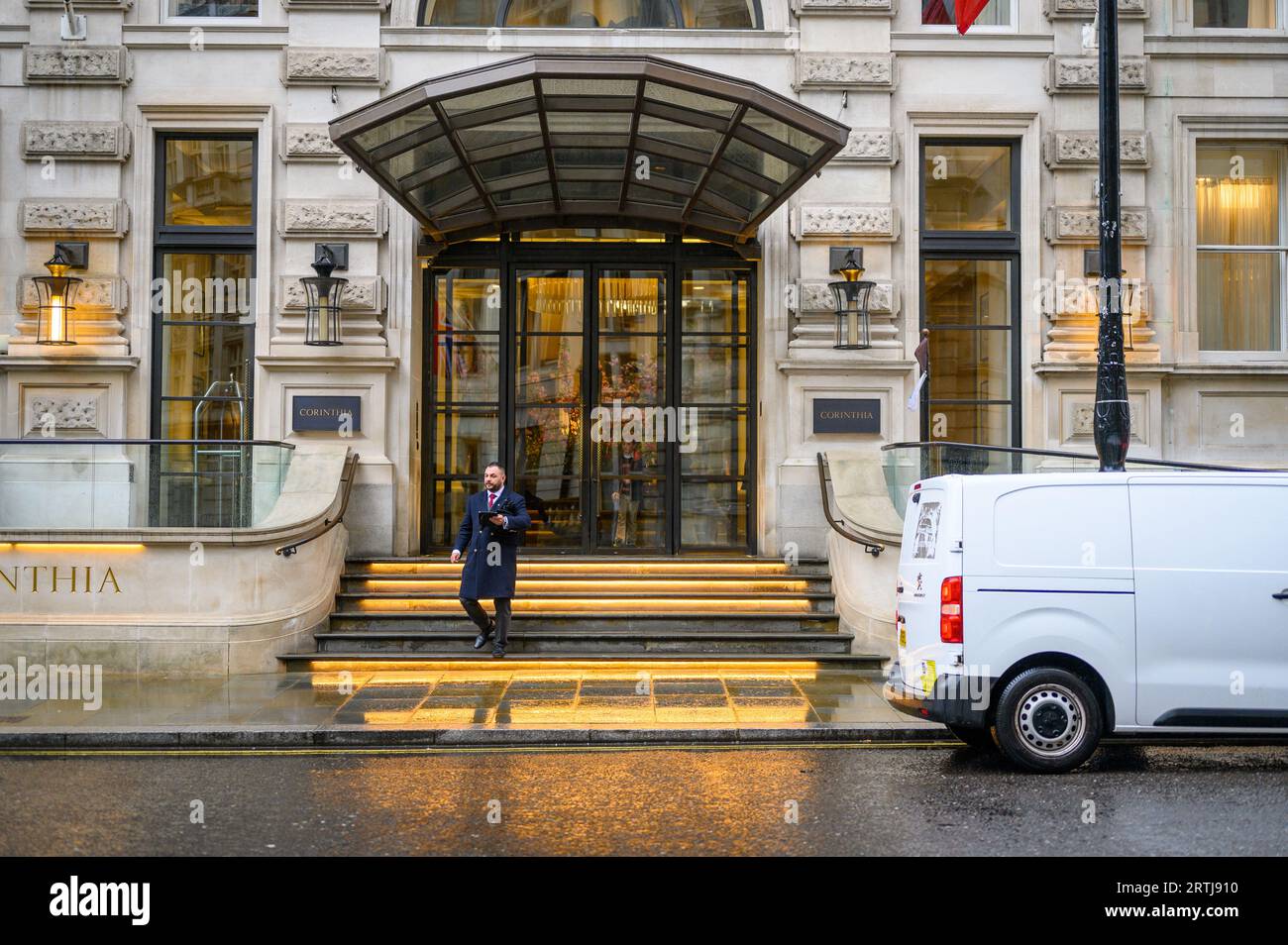 LONDON - 24. April 2023: Ein Türsteher steht auf den Stufen des Eingangs zum Corinthia Hotel und begrüßt Sie an einem regnerischen Tag im Herzen der ci Stockfoto