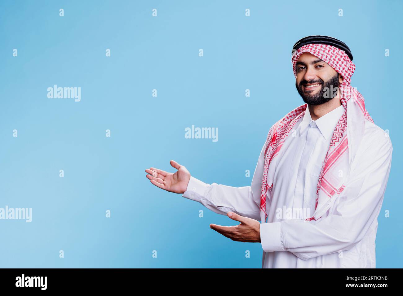 Muslimischer Mann mit traditionellem Gewand und Kopftuch, der sich mit den Händen zur Seite bewegt, während er für einen Studioschuss posiert. Arabischer Vertriebsmitarbeiter, der für Produkte wirbt, während er zeigt, dass er Platz mit Waffen kopiert Stockfoto