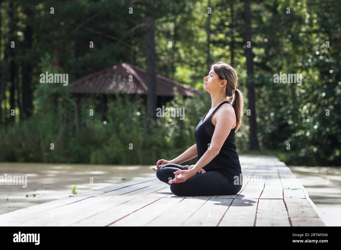 Eine Frau übt Meditation, sitzt in Lotusposition auf einer Holzbrücke in einem Park an einem sonnigen Sommermorgen und macht Yoga in schwarzer Sportkleidung Stockfoto