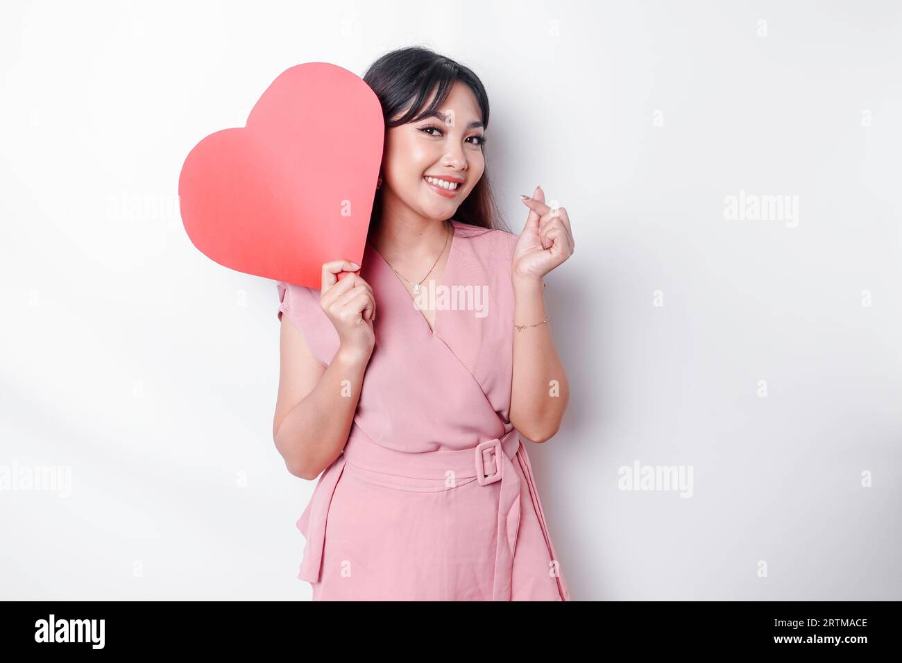 Eine glückliche junge asiatische Frau, die eine pinkfarbene Bluse trägt, fühlt sich wie eine romantische Geste an, die zarte Gefühle ausdrückt und ein rotes, herzförmiges Papier hält Stockfoto