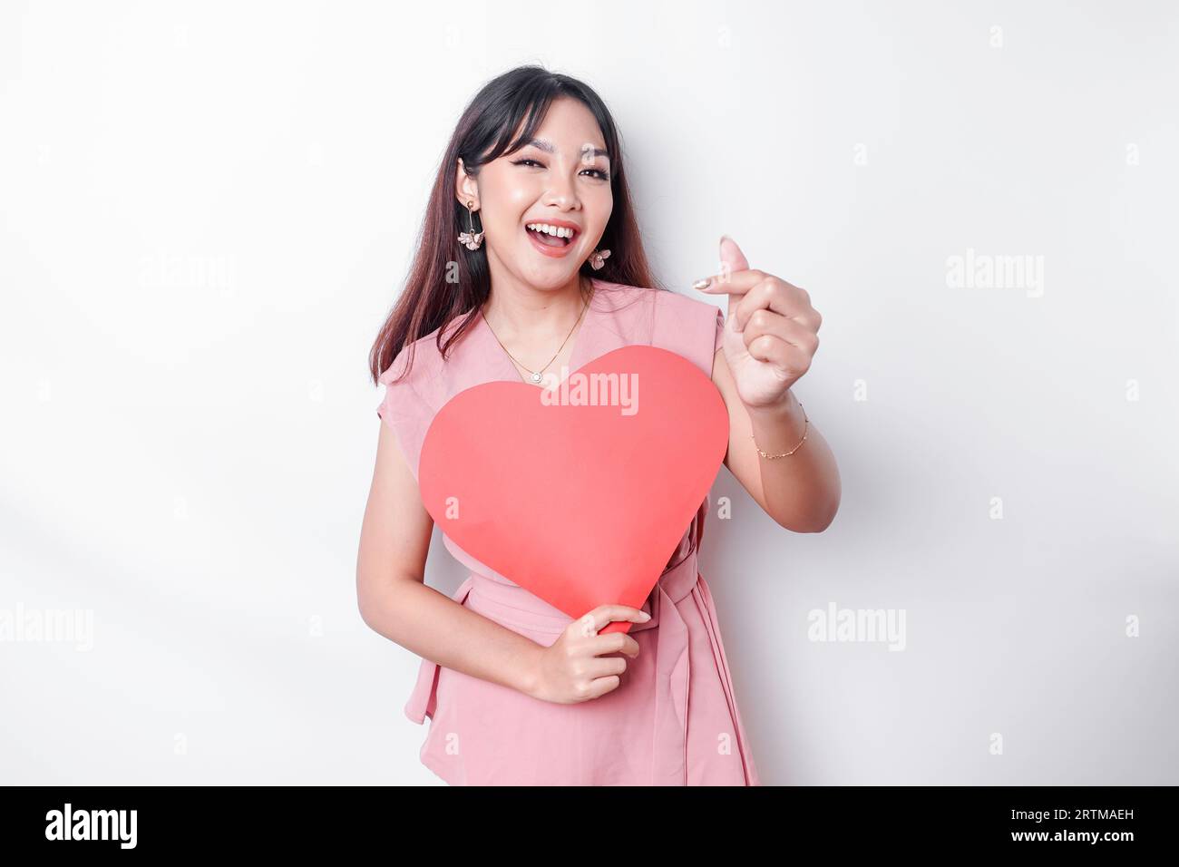 Eine glückliche junge asiatische Frau, die eine pinkfarbene Bluse trägt, fühlt sich wie eine romantische Geste an, die zarte Gefühle ausdrückt und ein rotes, herzförmiges Papier hält Stockfoto