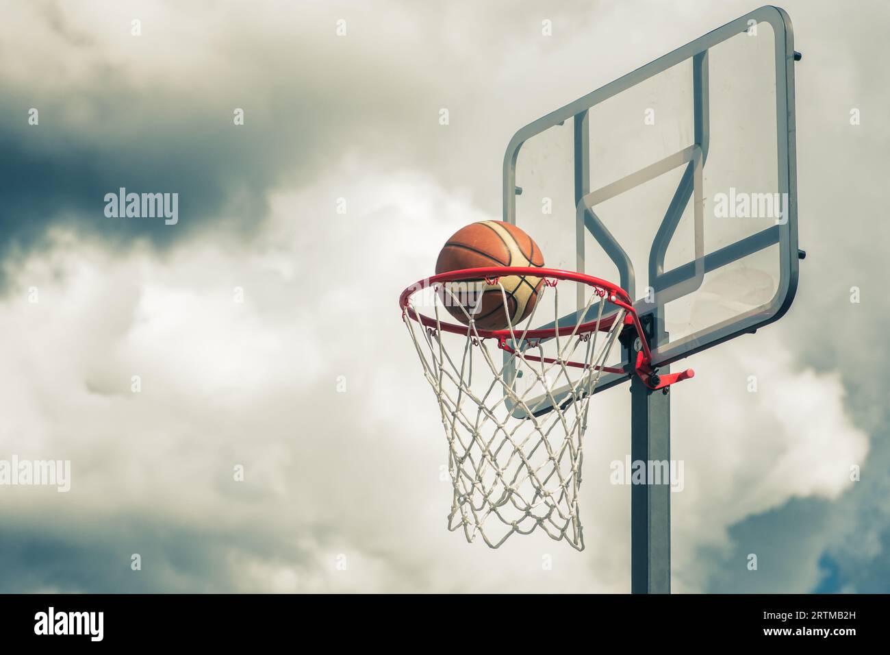 Der orangefarbene Basketballball tritt in den Basketballkorb ein. Spielen auf der Straße auf dem Outdoor-Court mit einem dramatischen Himmel und Wolken im Hintergrund, nah Stockfoto