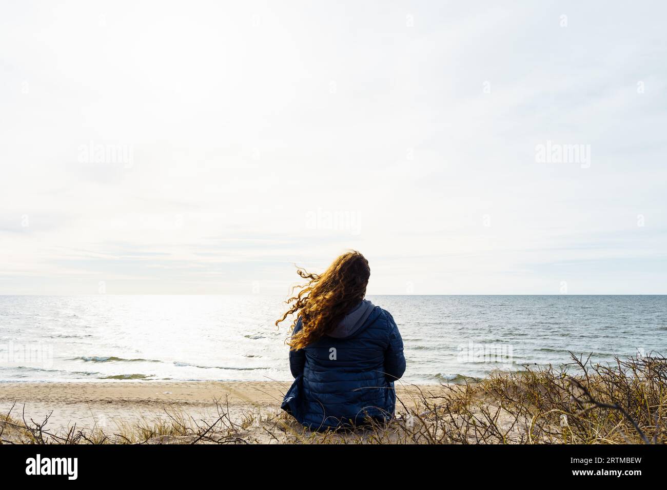 Lockige Frau in einer dünnen Daunenjacke, die abends am Meer sitzt, Haare flattern im Wind Stockfoto