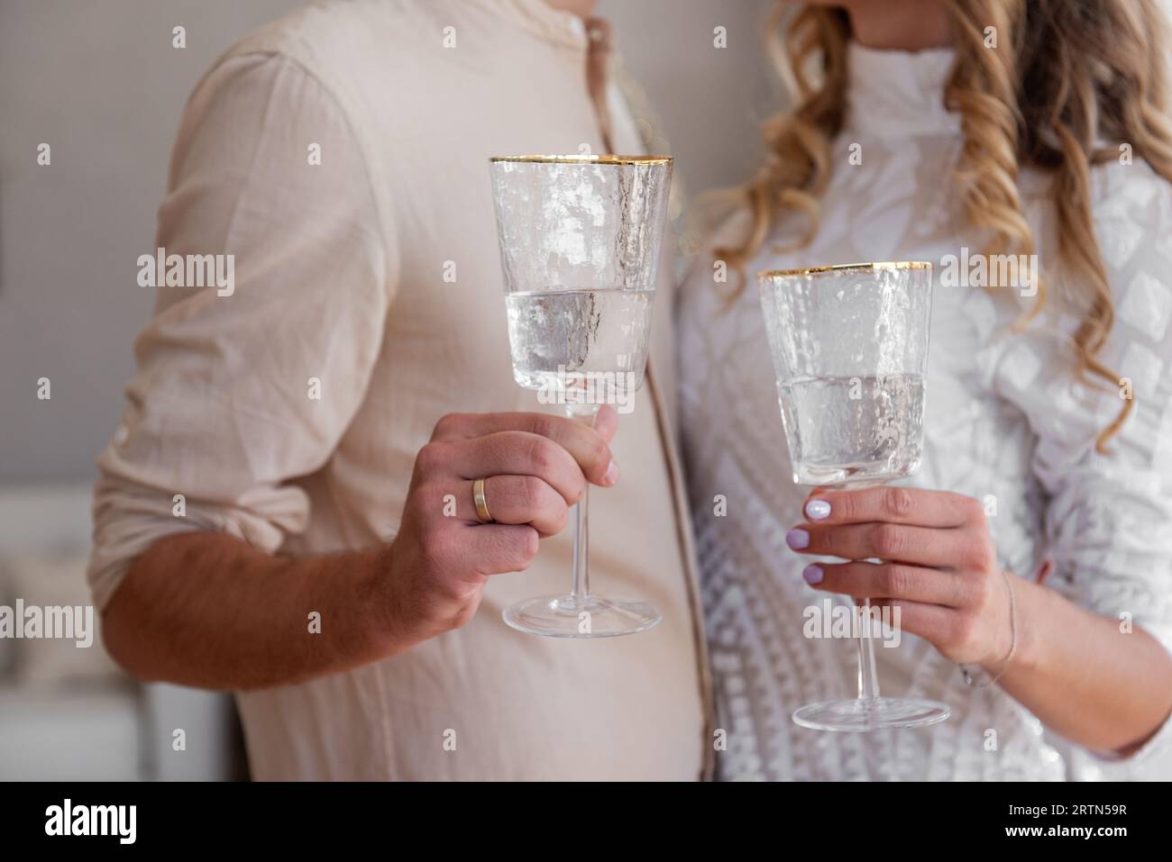Das Paar hält und toastet Champagnergläser. Nahaufnahme der Hände, die trendige Gläser mit Wein halten. Einfarbige beige Kleidung. Gemütlicher Stil. Liebhaber machen es Stockfoto