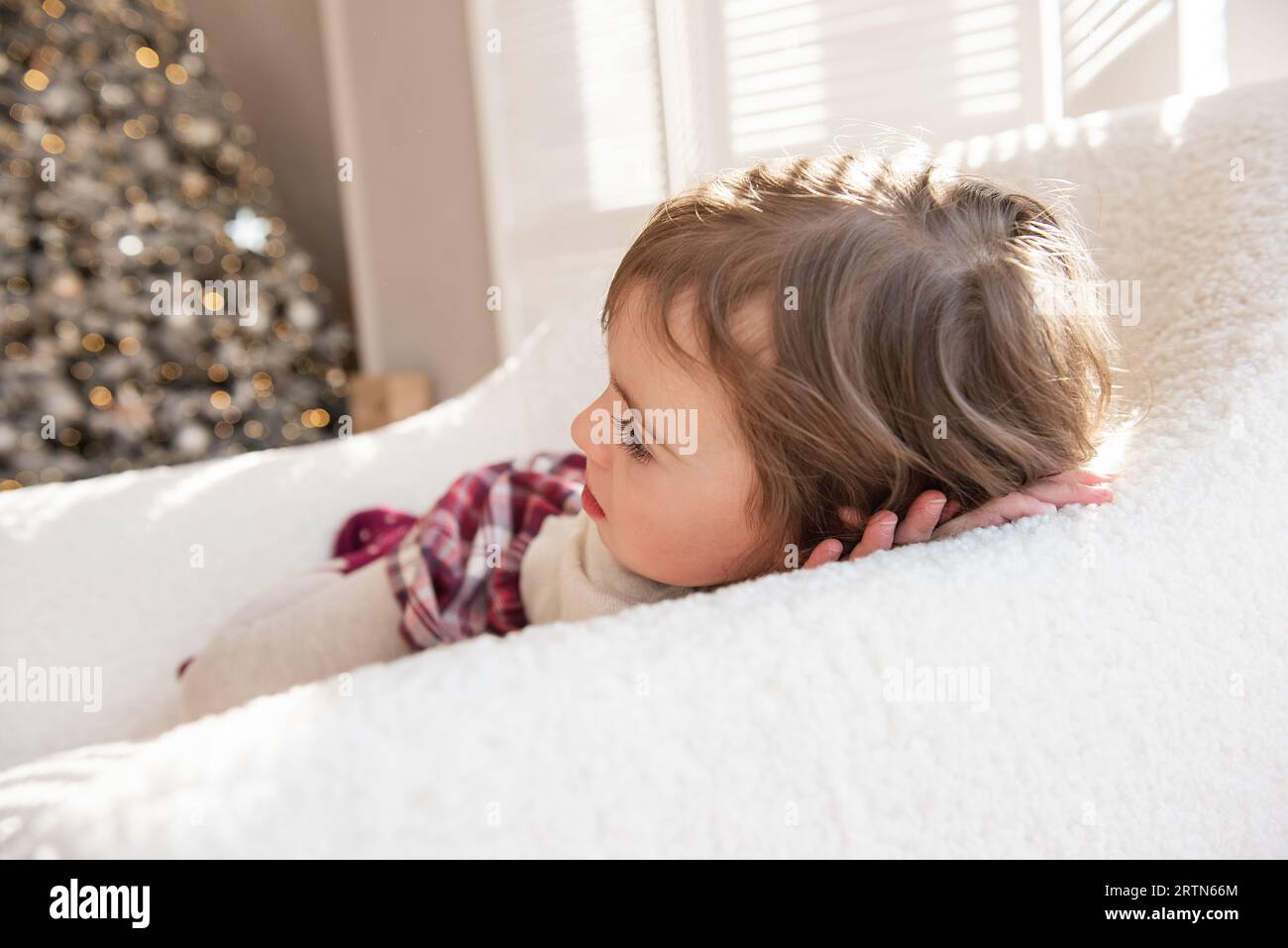 Ein nachdenkliches, träumendes kleines Mädchen liegt in einem weichen weißen Sessel und träumt von einem Geschenk des Weihnachtsmanns in der Nähe des Weihnachtsbaums. Kind in gemütlicher Atmosphäre im Stockfoto
