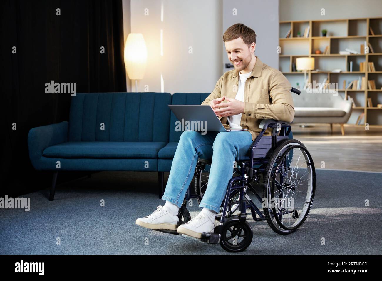 Gutaussehender Mann mit einer Behinderung, der im Rollstuhl sitzt, mit Laptop auf den Knien und die Kamera betrachtet Stockfoto