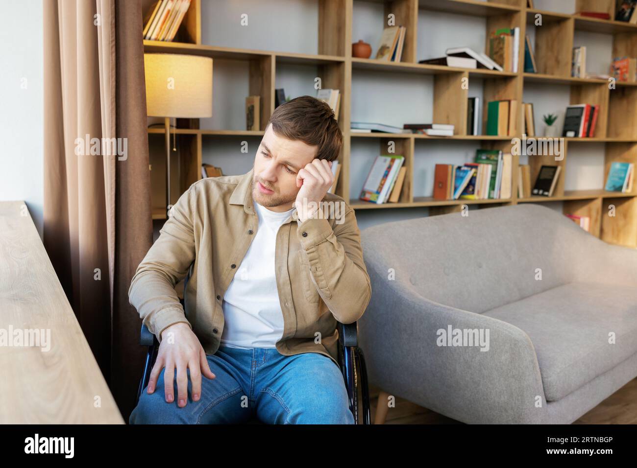 Ein junger, gutaussehender Mann im Rollstuhl blickt mit einem traurigen Blick aus dem Fenster. Stockfoto