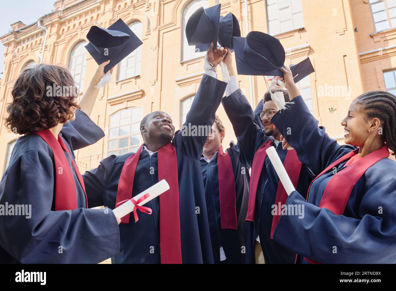 Eine multiethnische Gruppe von Studenten in Gewändern, die ihren Hut aufrichten und ihren Abschluss feiern Stockfoto