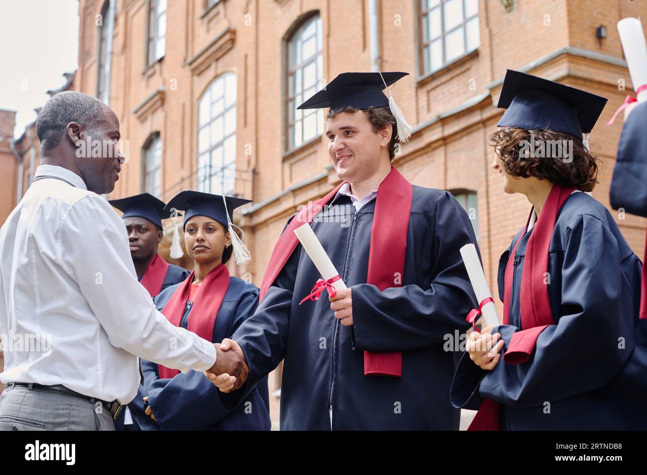 Der Lehrer schüttelt den Schülern die Hand und gratuliert ihnen zum Abschluss der Universität, während sie draußen stehen Stockfoto