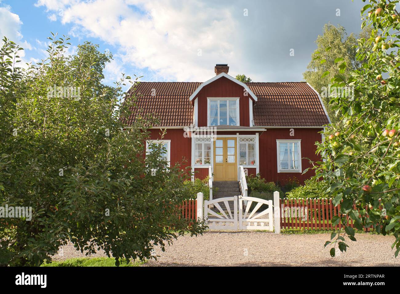 Ein typisches rot-weißes schwedisches Haus im Kleinen. Weißes Gartentor, brauner Zaun. Grüner Rasen im Garten. Bäume im Vordergrund. Blauer Himmel mit sm Stockfoto
