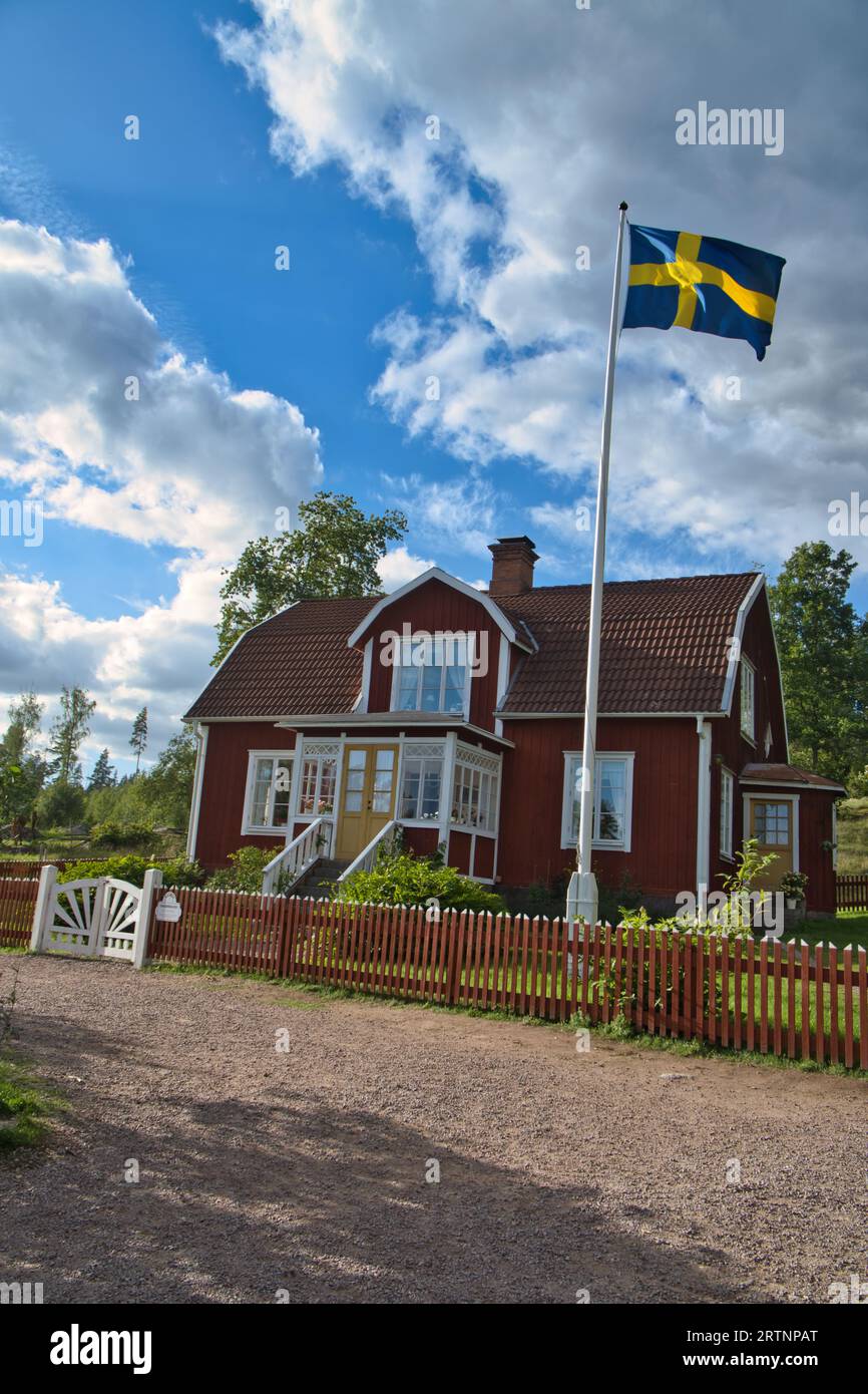 Ein typisches rot-weißes schwedisches Haus im Kleinen. Weißes Gartentor, brauner Zaun. Fahnenmast mit schwedischer Flagge. Grüner Rasen im Garten. Bäume im Hintergrund Stockfoto