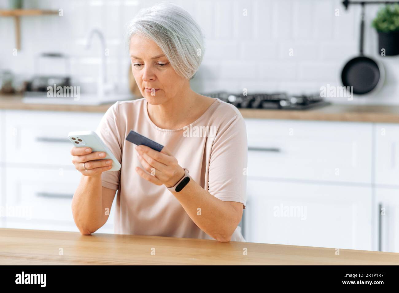 Konzentrierte weiße, reife, grauhaarige Frau, sitzt zu Hause in einer Küche, hält ein Smartphone und eine Kreditkarte, macht eine Online-Bestellung, bezahlt Einkäufe oder Rechnungen mit einer Bankkarte und einem Antrag Stockfoto