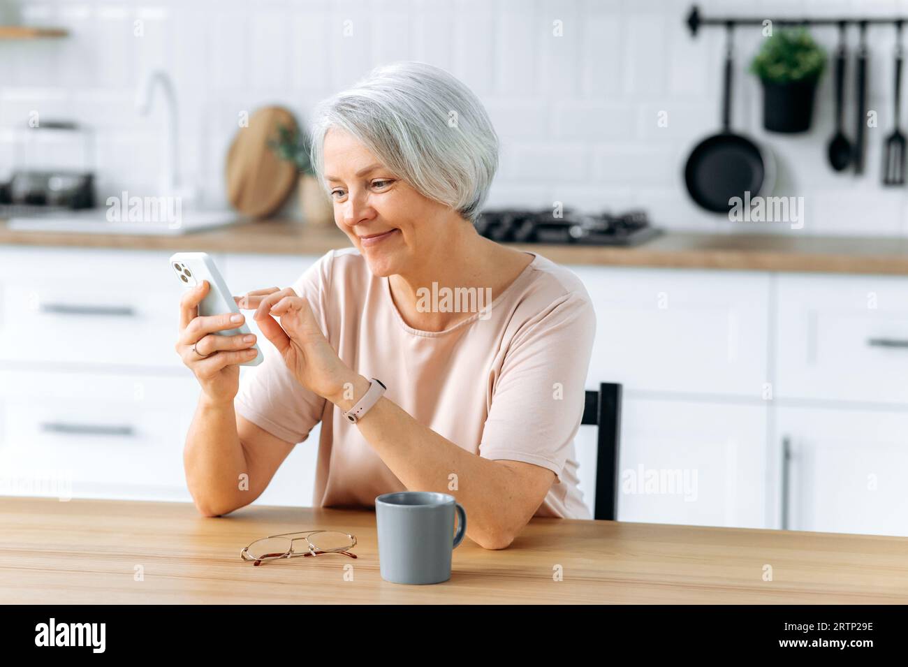 Glückliche, hübsche, weiße, grauhaarige Frau mittleren Alters, sitzt zu Hause in der Küche, hält ihr Smartphone in der Hand, benutzt es, um in sozialen Netzwerken zu kommunizieren, Nachrichten zu lesen, macht eine Online-Bestellung, lächelt Stockfoto